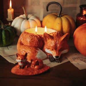 Puede incluir: Un portavelas con forma de zorro con dos mechas encendidas, que muestra un zorro adulto durmiendo y una figura de un zorro pequeño delante. El portavelas está rodeado por una calabaza verde, una blanca, una naranja y una vela encendida.