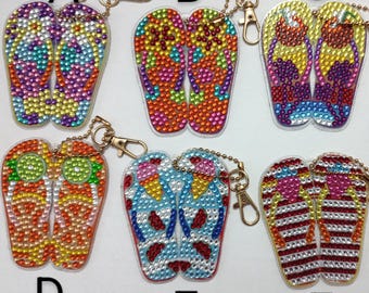 May include: A collection of seven beaded flip-flop keychains in various colors. Each keychain features a pair of flip-flops adorned with colorful beads, attached to a chain and a clasp. The keychains are in shades of purple, orange, blue, and red.