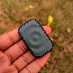 May include: A dark green, rectangular pendant with a circular design in the center, set within a black frame. The pendant is held in a hand against a blurred background of grass and foliage.