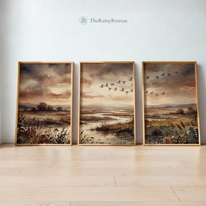 Op de afbeelding: Drie ingelijste aquarel landschap prints. Elk toont een moeraslandschap in gedempte bruin-, beige- en grijstinten. Eén print toont een vlucht vogels. De frames zijn goudkleurig en het kunstwerk wordt gepresenteerd op een lichte houten vloer.