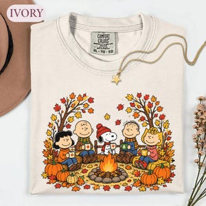 May include: An ivory Comfort Colors t-shirt featuring a graphic of the Peanuts characters sitting around a campfire in an autumn forest. The design includes colorful fall leaves, pumpkins, and the characters holding mugs. A gold star necklace is included.