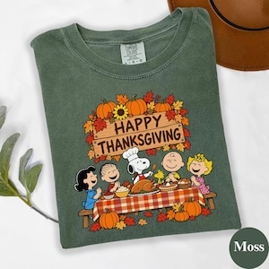 May include: A moss green crew neck t-shirt featuring a graphic of the Peanuts characters at a Thanksgiving dinner table. The text reads Happy Thanksgiving above the characters, with pumpkins, autumn leaves, and a turkey on the table.