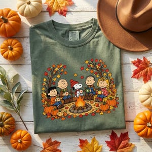 May include: An olive green t-shirt featuring a graphic print of the Peanuts characters sitting around a campfire with autumn leaves and pumpkins. The shirt has a crew neckline and a Comfort Colors label printed on the inside of the collar.