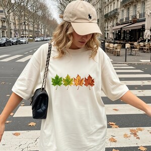 May include: An off-white short-sleeve t-shirt featuring a horizontal row of four maple leaves in a color gradient from green to yellow to orange and brown. The shirt is paired with a beige baseball cap and a black quilted shoulder bag.