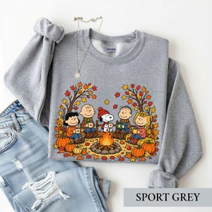 May include: A light gray crewneck sweatshirt featuring a graphic of the Peanuts characters sitting around a campfire with autumn leaves and pumpkins. The design includes Snoopy, Charlie Brown, and friends holding mugs. A thin silver necklace is included.
