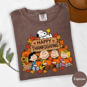 May include: A brown crew neck t-shirt featuring a graphic of Snoopy, Woodstock, and the Peanuts gang with pumpkins and autumn leaves. The text reads Happy Thanksgiving on a wooden sign. The shirt has a Comfort Colors label in the neck.
