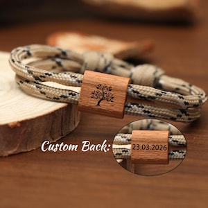 Puede incluir: Una pulsera tejida con un cierre de madera con un diseño de árbol. La pulsera está hecha de cuerda beige y negra. El cierre está grabado con un árbol y el texto "Custom Back:" es visible. Una imagen más pequeña muestra la fecha "23.03.2026" en el cierre.