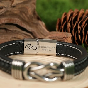 May include: Black leather bracelet with white stitching and a silver clasp. The clasp has the text "Believe in you like I do" and an infinity symbol with a heart. The bracelet is on a wooden surface.