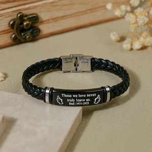 Könnte beinhalten: Ein schwarzes geflochtenes Lederarmband mit einer zentralen schwarzen Metallplatte, die mit dem Text "Those we love never truly leave us. Dad | 1931-2025" und zwei Engelsflügel-Symbolen graviert ist. Das Armband hat einen silberfarbenen Metallverschluss.