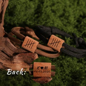May include: Two personalized bracelets with wooden accents. One bracelet features a brown cord and the other a black cord. Each has a wooden rectangular piece with etched lines. The back of one bracelet shows the initials "K♥M" and the date "02.03.2020".