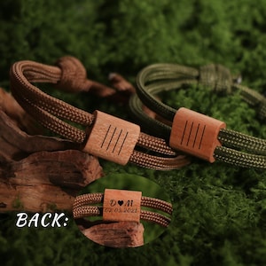 May include: Two bracelets with wooden accents. One bracelet is brown, the other is olive green. Each bracelet has a wooden accent with four vertical lines. The back of one bracelet shows initials and a date.
