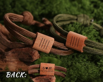 May include: Two bracelets with wooden accents. One bracelet is brown, the other is olive green. Each bracelet has a wooden accent with four vertical lines. The back of one bracelet shows initials and a date.