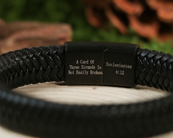 May include: Black braided leather bracelet with a magnetic clasp. The clasp is engraved with the text "A Cord Of Three Strands Is Not Easily Broken" and "Ecclesiastes 4:12". The bracelet is a dark, neutral color.