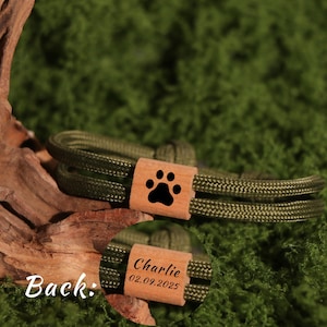 May include: Olive green dog collar with a wooden accent featuring a black paw print. The back of the wooden accent is engraved with the name "Charlie" and the date "02.09.2025". The collar is made of a braided rope material.
