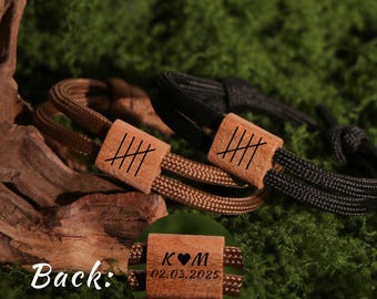 May include: Two personalized bracelets with wooden accents. One bracelet features a brown cord and the other a black cord. Each has a wooden rectangular piece with etched lines. The back of one bracelet shows initials and a date.
