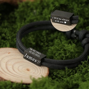 Op de afbeelding: Zwart gevlochten armband met een rechthoekige zwarte metalen kraal gegraveerd met de naam "James", data en een hartsymbool. Een andere kraal leest "I Will be always with you". De armband ligt op een houten schijf.