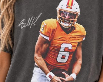 Puede incluir: Una camiseta de manga corta gris carbón con el gráfico de un jugador de fútbol americano con una camiseta naranja con el número 6 y un casco blanco. La camiseta incluye una firma impresa y el texto PEPPER dentro de un círculo.