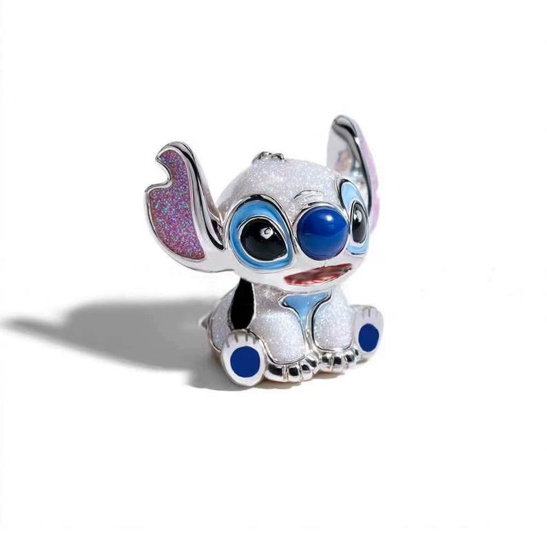 May include: A small, glittery white figurine of the character Stitch, featuring large ears with pink glitter interiors, dark blue eyes, a round blue nose, and blue accents on the feet. The character is depicted in a seated, forward-facing pose.
