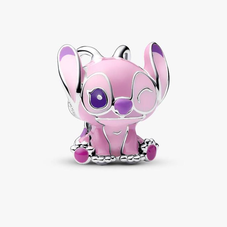 Puede incluir: Un charm de plata con forma de personaje que gui&ntilde;a un ojo con orejas grandes, con detalles de esmalte rosa claro en el cuerpo y las orejas, un ojo morado oscuro y una nariz morada. El personaje est&aacute; en posici&oacute;n sentada con almohadillas visibles.