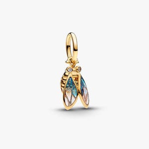 Puede incluir: Un charm dorado con forma de insecto estilizado, con alas esmaltadas en azul, blanco y dorado. El charm tiene un lazo para sujetarlo a una pulsera o collar y está adornado con pequeñas piedras preciosas transparentes.