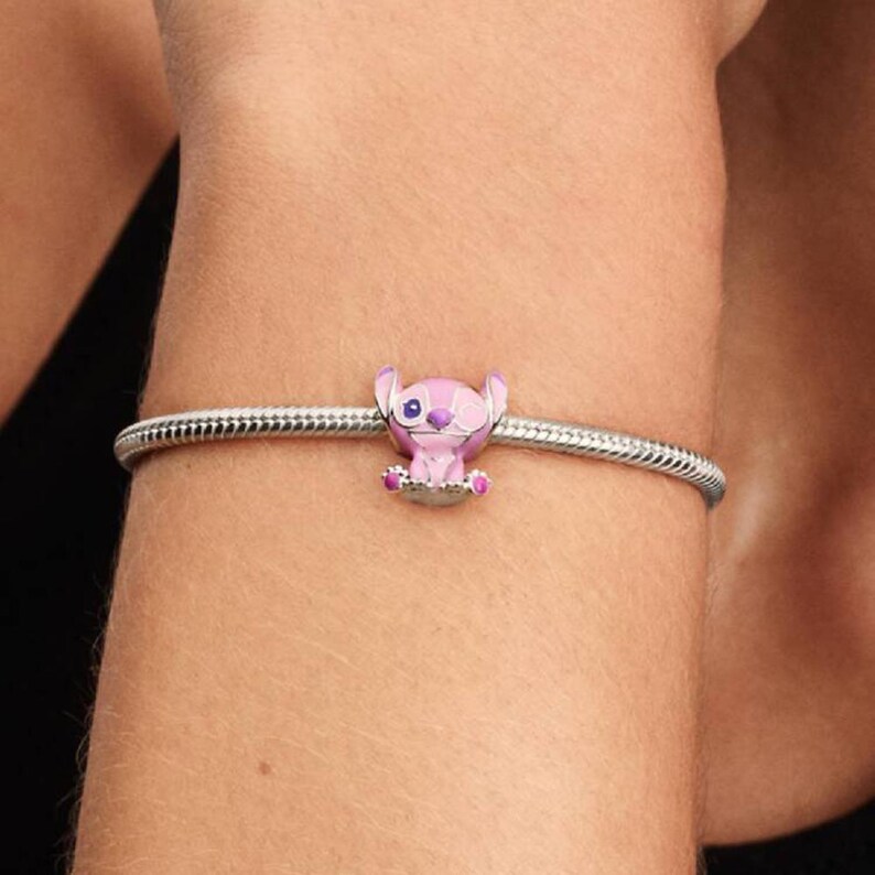 Puede incluir: Una pulsera de cadena de serpiente plateada con un charm con la forma del personaje Stitch, realizado en esmalte rosa claro con detalles en rosa oscuro en las orejas y patas, y una peque&ntilde;a nariz morada, centrado en la banda met&aacute;lica.