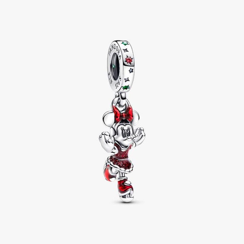 May include: A silver charm featuring Minnie Mouse in a red dress and bow. The charm has a dangling design with a ring at the top, adorned with stars and the brand name. The Minnie Mouse figure is detailed with red enamel.
