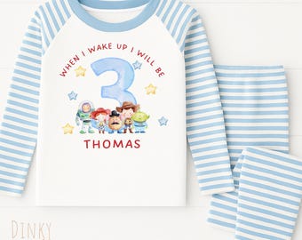 May include: A light blue and white striped pajama set. The top has a white front with the text "WHEN I WAKE UP I WILL BE 3 THOMAS" and a graphic of Toy Story characters. The sleeves are light blue and white striped.