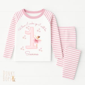 May include: A two-piece pyjama set with a white top and pink striped trousers. The white top features a pink number one, a ballerina, and the text "When I wake up I will be Sienna."