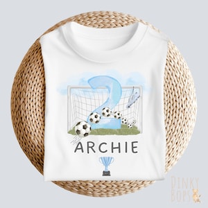 May include: White t-shirt with a soccer-themed design for a two-year-old. The design includes a large blue number 2, soccer balls, a goal, and the name "ARCHIE" in black. A trophy is also included in the design.