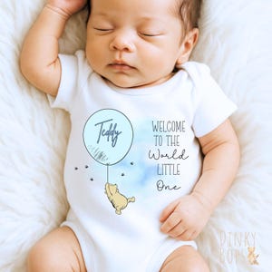 May include: A white baby bodysuit featuring a light blue balloon graphic with the name "Teddy". A cartoon bear hangs from the balloon. The text "WELCOME TO THE World LITTLE One" is printed on the garment.