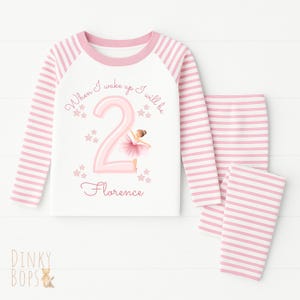 May include: A two-piece pajama set. The white top has pink striped sleeves and the pants are pink striped. The top features the text "When I wake up I will be 2 Florence" with a ballerina graphic. The pajamas are designed for a child.