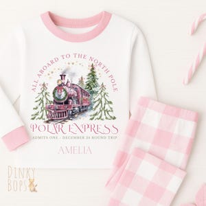 May include: A white pajama set with pink trim and pink and white checked trousers. The top displays a watercolour illustration of a red train, with the text "POLAR EXPRESS" and "ALL ABOARD TO THE NORTH POLE".