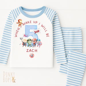 May include: A two-piece pajama set with a white top and blue striped sleeves, plus matching trousers. The top features a blue number 5 with superhero graphics and the text "When I wake up I will be" and the name "Zach".