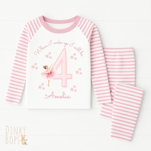 May include: A pink and white striped pajama set. The top has a white body with pink sleeves and a pink neckline. It features a ballerina graphic, the number 4, and the name "Amelia". The text "When I wake up I will be" is also printed on the top. The set includes matching striped trousers.