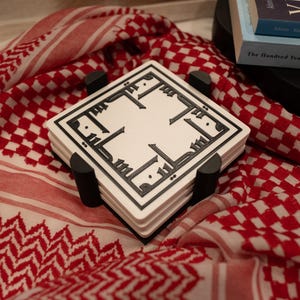 May include: A stack of square white coasters with black Arabic calligraphy, held in a black holder. The coasters rest on a red and white checkered scarf. Books are visible in the background.