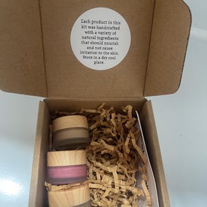 May include: An open cardboard box containing three small jars with wooden lids. The jars are filled with different colored products, including a pink and two shades of brown. The box also contains shredded brown paper and a white label with text.
