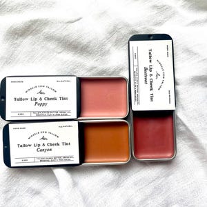 K&ouml;nnte beinhalten: Vier kleine, rechteckige Dosen mit Miracle Cow Tallow Lippen- und Wangenfarben. Die Farbt&ouml;ne sind in Korallen-, Orange- und Rott&ouml;nen gehalten. Jede Dose ist mit dem Produktnamen und den Inhaltsstoffen beschriftet. Die Farbt&ouml;ne Poppy, Canyon und Beetroot sind sichtbar.