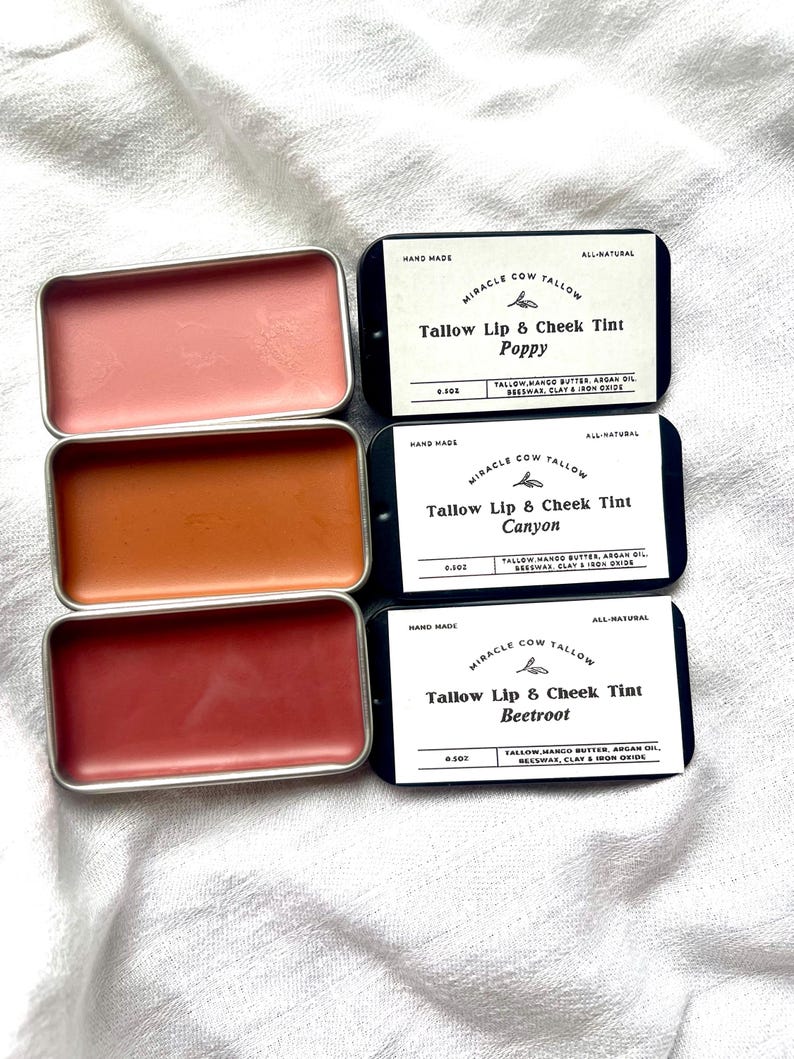 K&ouml;nnte beinhalten: Drei kleine, rechteckige Dosen mit Miracle Cow Tallow Lippen- und Wangenfarbe in den Farbt&ouml;nen Poppy, Canyon und Beetroot. Jede Dose befindet sich in einem silbernen Beh&auml;lter mit einem schwarzen Etikett. Die Etiketten listen die Inhaltsstoffe und die W&ouml;rter "Hand Made" und "All-Natural" auf.