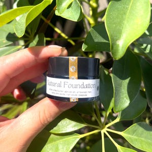 May include: A small, black jar of "Natural Foundation" held in a hand, with a white label and gold band featuring daisy accents. The label lists ingredients like shea butter and apricot oil. Green leaves form the background.