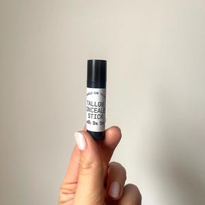 May include: A black concealer stick with a white label. The label reads "Tallow Concealer Stick with tea tree". The product is held in a hand against a plain background. The concealer stick is approximately 3 inches long.