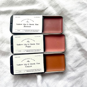 K&ouml;nnte beinhalten: Drei kleine, rechteckige Dosen Miracle Cow Tallow Lip & Cheek Tint in den Farbt&ouml;nen Beetroot, Poppy und Canyon. Jede Dose hat ein schwarzes Etikett mit wei&szlig;em Text. Die Farbt&ouml;ne sind verschiedene Rot- und Oranget&ouml;ne. Der Hintergrund ist ein wei&szlig;er, strukturierter Stoff.