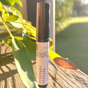 May include: A black mascara tube with a pink label that reads "Rosemary Mascara." The tube is set against a backdrop of green leaves and a wooden surface, with the text "FACEBOOK" visible on the side.