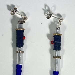 Puede incluir: Un par de pendientes colgantes con postes plateados. Cada pendiente presenta un tubo transparente envuelto con hilo azul oscuro y acentuado con anillos dorados y una peque&ntilde;a cuenta roja. La parte inferior de cada pendiente tiene una cadena de cuentas azules.