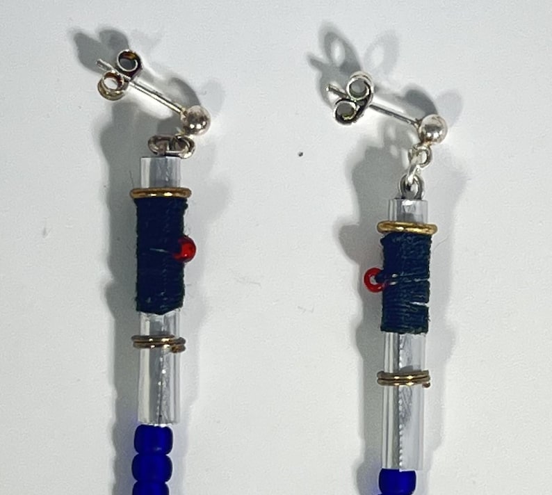 Puede incluir: Un par de pendientes colgantes con postes plateados. Cada pendiente presenta un tubo transparente envuelto con hilo azul oscuro y acentuado con anillos dorados y una peque&ntilde;a cuenta roja. La parte inferior de cada pendiente tiene una cadena de cuentas azules.