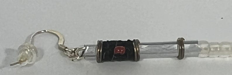 Puede incluir: Primer plano de un pendiente plateado con un cierre de pl&aacute;stico transparente. El pendiente presenta una cuenta cil&iacute;ndrica negra con un acento rojo y cuentas rectangulares transparentes.