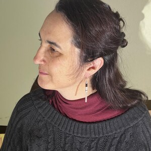 Puede incluir: Una mujer con un su&eacute;ter de punto trenzado gris oscuro y un cuello alto burdeos. Lleva un par de pendientes largos y colgantes con detalles en negro, blanco y plateado. Los pendientes son de forma rectangular.