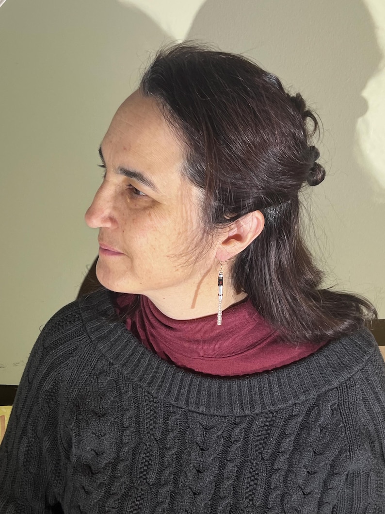 Puede incluir: Una mujer con un su&eacute;ter de punto trenzado gris oscuro y un cuello alto burdeos. Lleva un par de pendientes largos y colgantes con detalles en negro, blanco y plateado. Los pendientes son de forma rectangular.