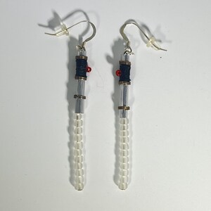 Puede incluir: Un par de pendientes hechos a mano. Cada pendiente presenta un tubo cil&iacute;ndrico transparente con cuentas blancas en la parte inferior. La secci&oacute;n superior tiene una secci&oacute;n envuelta en azul oscuro, una peque&ntilde;a cuenta roja y un gancho plateado.