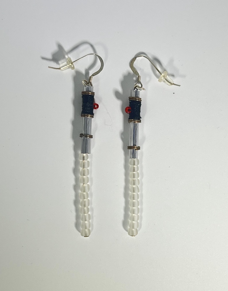 Puede incluir: Un par de pendientes hechos a mano. Cada pendiente presenta un tubo cil&iacute;ndrico transparente con cuentas blancas en la parte inferior. La secci&oacute;n superior tiene una secci&oacute;n envuelta en azul oscuro, una peque&ntilde;a cuenta roja y un gancho plateado.