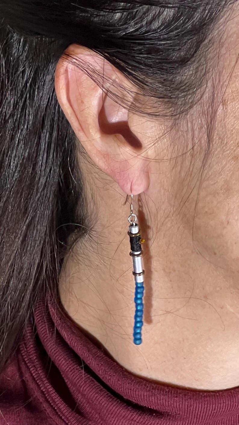 Puede incluir: Un solo pendiente con dise&ntilde;o de sable de luz. El pendiente presenta una empu&ntilde;adura de metal plateado con detalles en negro y dorado, y una hoja de cuentas azules. El pendiente est&aacute; suspendido de un gancho plateado.